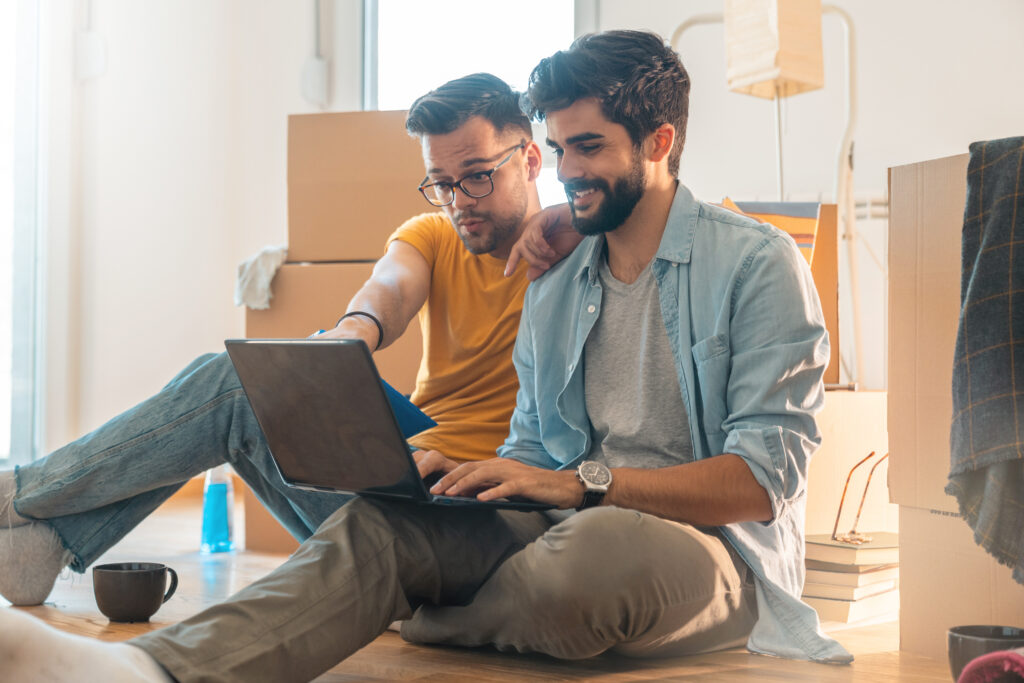 Zwei Männer sitzen lächelnd auf dem Boden und schauen gemeinsam auf einen Laptop